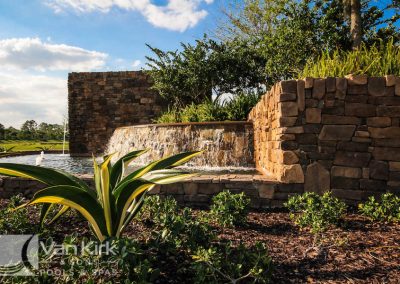 Stone waterfall feature in a landscaped garden with lush greenery and clear blue sky.