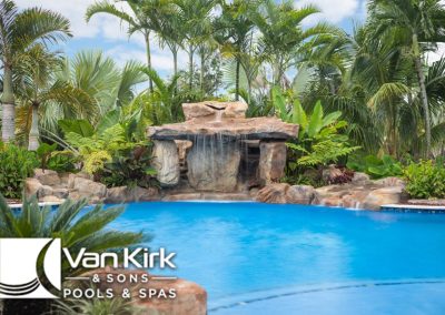 Tropical swimming pool with waterfall feature, surrounded by lush greenery, by Van Kirk & Sons Pools & Spas.