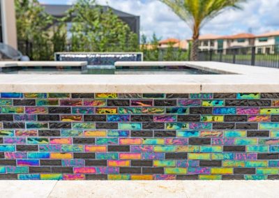 Colorful iridescent tiles surrounding a modern outdoor pool with blurred greenery and buildings in the background.