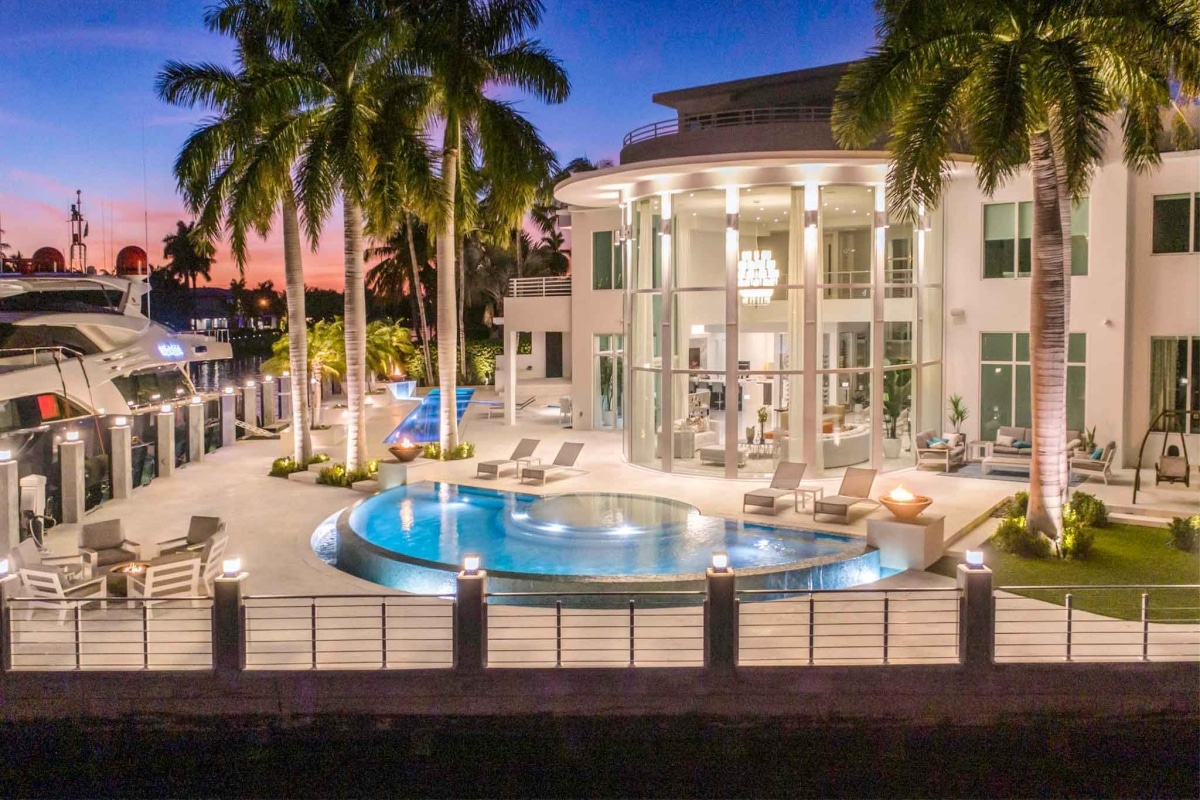 Luxurious waterfront mansion at sunset with a pool, palm trees, and a yacht docked nearby.