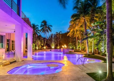 Luxurious tropical freeform pool area with vibrant evening lighting and palm trees surrounding a modern villa.