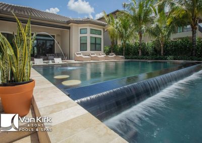 Modern infinity pool with waterfall feature, surrounded by tropical plants and sun loungers for relaxation.