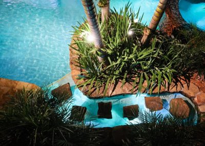 Aerial view of a tropical pool with stepping stones through a pool and lush palm trees, illuminated by vibrant lighting.