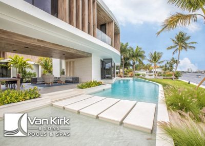 Modern luxury home with elegant pool and waterfront view, surrounded by palm trees. Step decks. Van Kirk & Sons Pools & Spas logo.