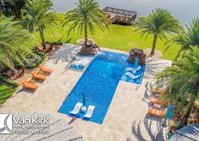 Aerial view of a luxurious straight line pool with palm trees and lounge chairs by Van Kirk & Sons Pools & Spas near a lake.
