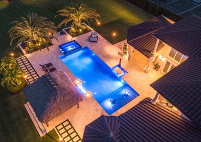 Aerial view of a lit backyard rectangular pool with palm trees, patio, and tiki hut at night.
