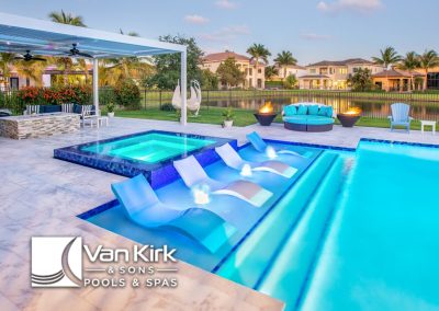 Luxurious backyard pool and spa with bubblers, modern patio furniture, palm trees, and serene lakeside view.
