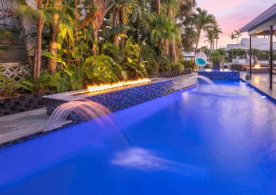 Luxurious backyard pool with water features, deck jets, fire ambiance, and tropical plants at sunset.