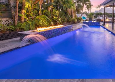 Luxurious backyard pool with waterfalls, fire feature, and tropical landscaping at sunset.