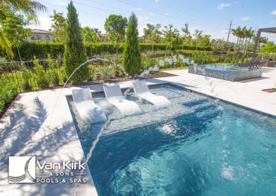 Luxurious backyard pool with laminar jets and lounge chairs, surrounded by greenery under a clear blue sky.