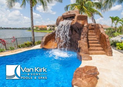 Luxury pool with rock waterfall by a scenic lake, designed by Van Kirk & Sons Pools & Spas, surrounded by palm trees.