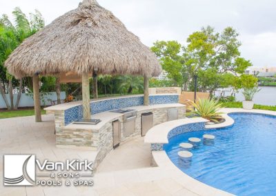 Poolside view with tiki hut, outdoor kitchen, and tropical landscaping by the water.