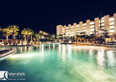 Luxurious hotel pool at night, glowing under lights with palm trees and building in the background.