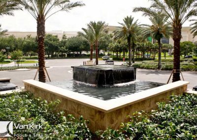 Elegant square fountain surrounded by palm trees and lush greenery in a landscaped area.