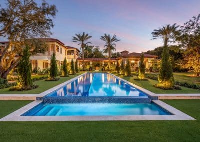 Luxurious villa with a long, illuminated swimming wet edge pool, surrounded by palm trees and greenery at sunset.