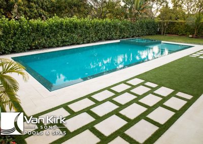 Modern backyard infinity edge pool with sleek design, lush greenery, and stylish paved area by Van Kirk & Sons Pools & Spas.