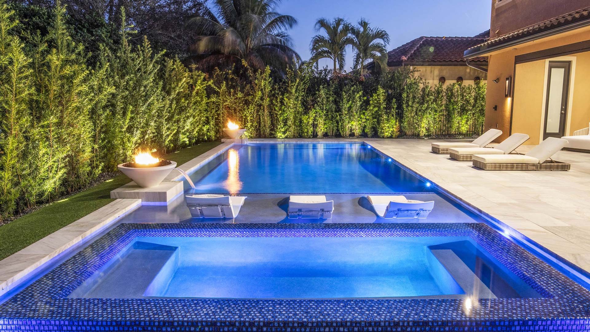 Luxurious Fort Lauderdale, FL backyard pool with glowing fire pits, surrounded by lush greenery and stylish lounge chairs at sunset.