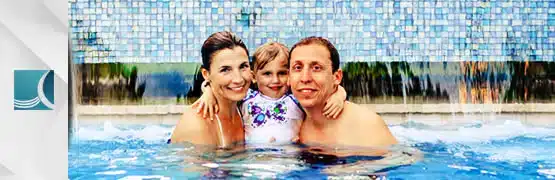 Family enjoying a swim in a pool with waterfall background, smiling and embracing in sparkling blue water.