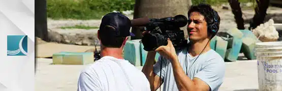 Cameraman filming a person outdoors, wearing a headset, with green foliage in the background.