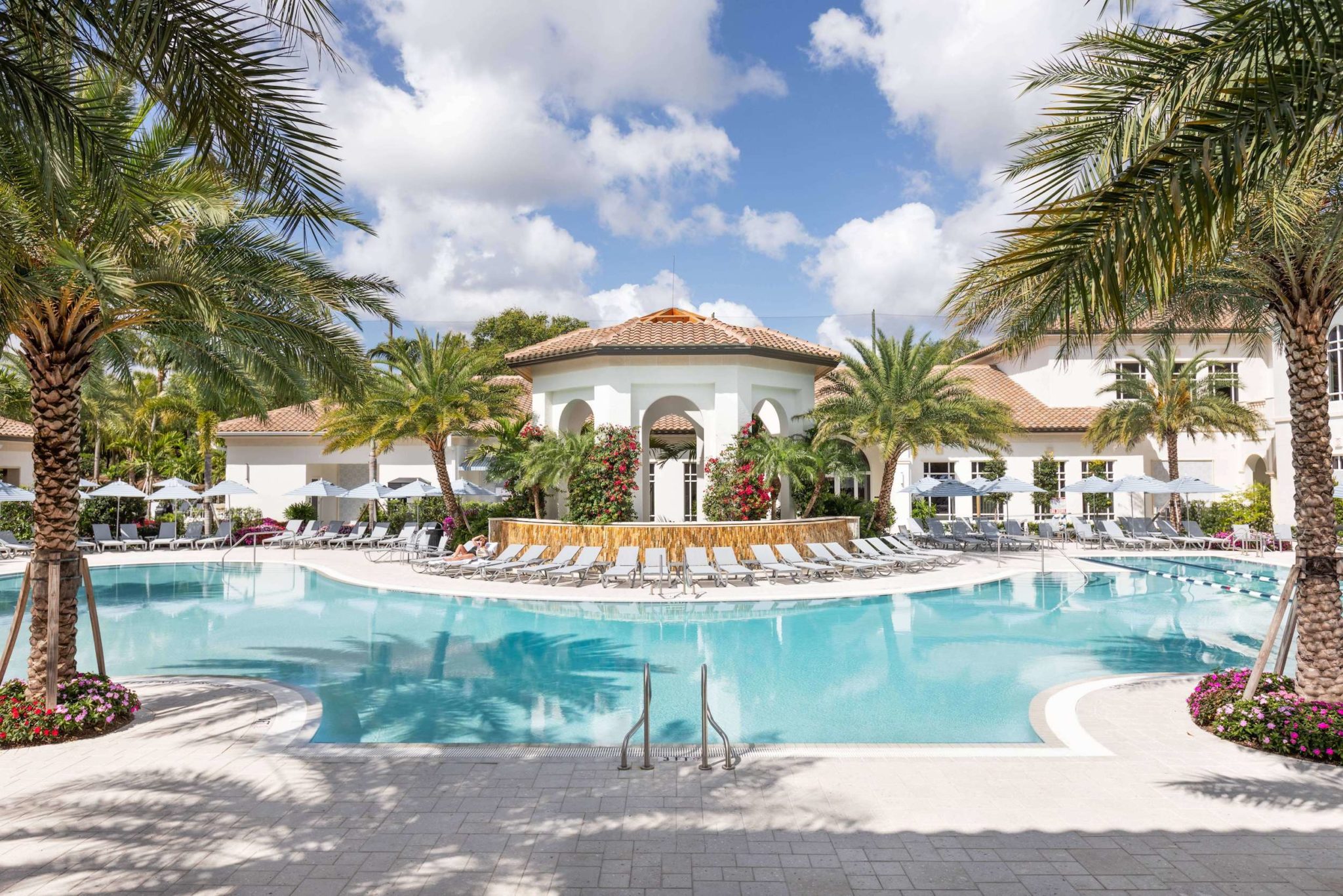 Luxurious resort pool surrounded by palm trees, sun loungers, and a Mediterranean-style building under a blue sky. in lake worth