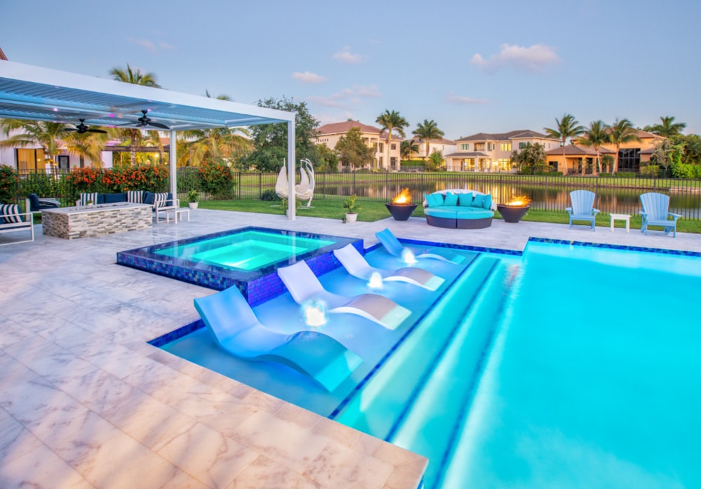 Luxury outdoor pool area with loungers, fire pits, and patio seating set against a serene residential backdrop.