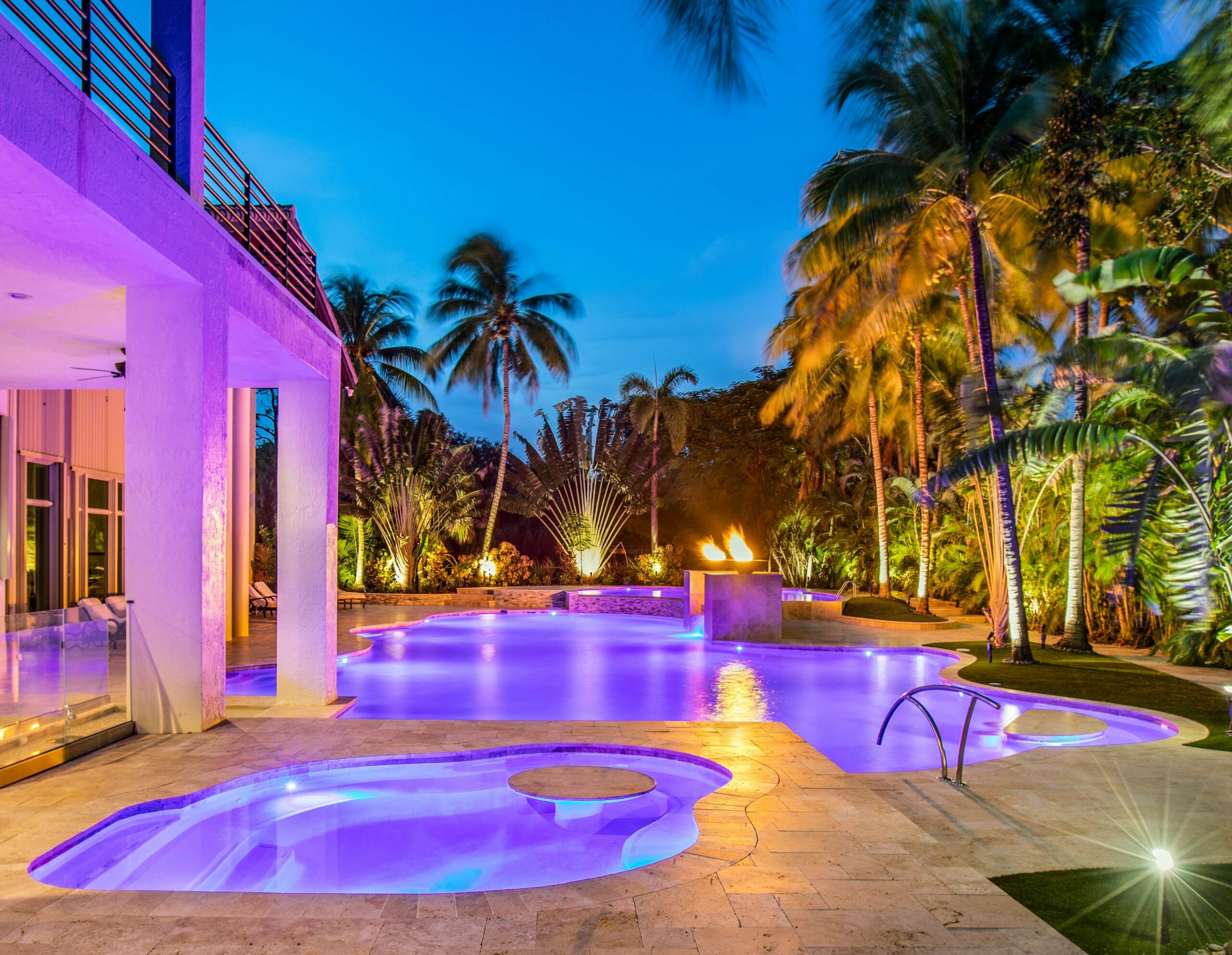 Luxury resort pool at night with vibrant lighting, surrounded by palm trees and tropical foliage in palm beach