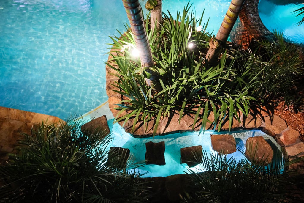 Tropical pool with lush greenery, palm trees, and stepping stones under ambient lighting, creating a serene evening atmosphere in plantation