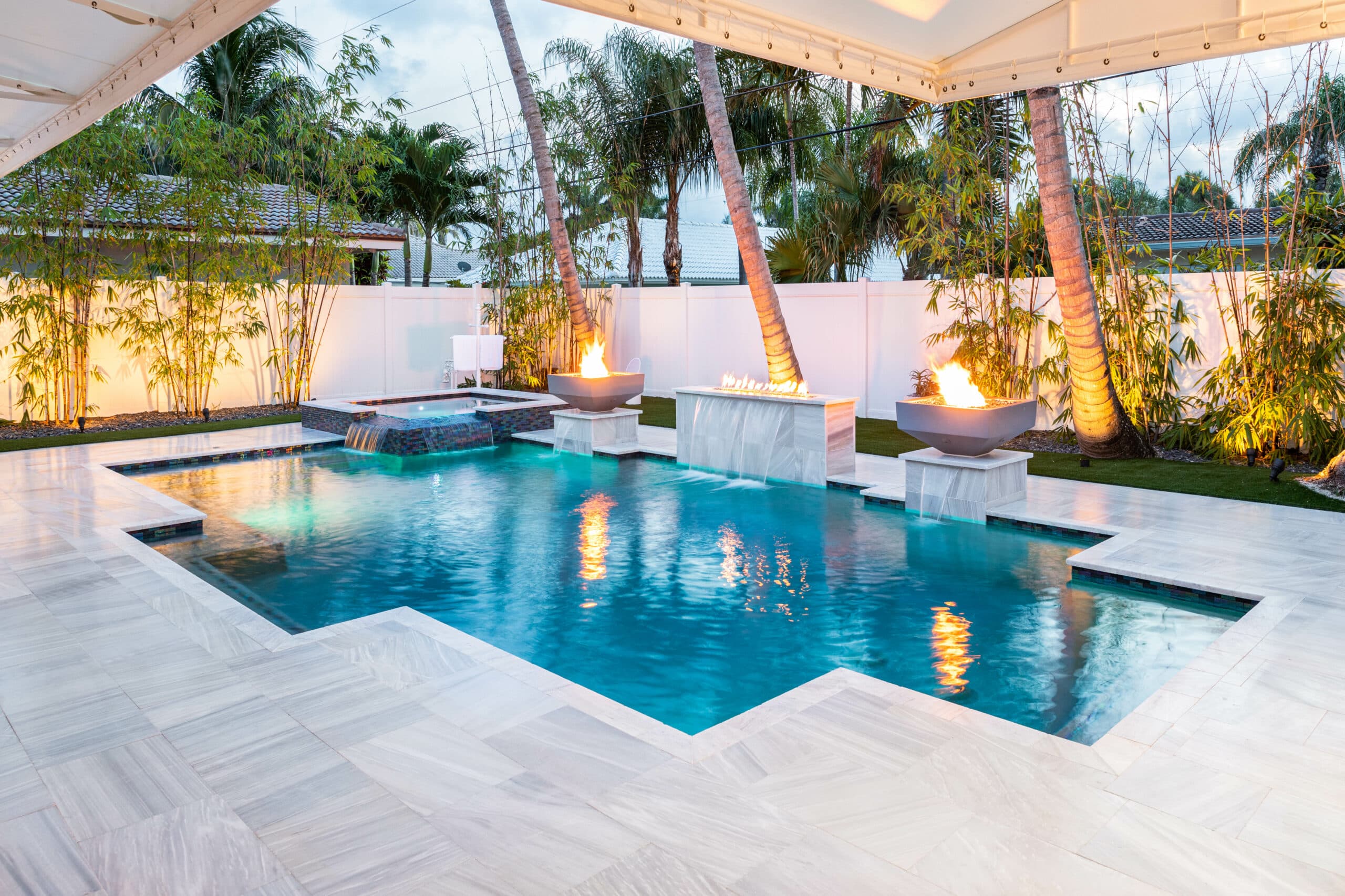 Luxurious outdoor pool in Delray Beach, Florida with palm trees and fire features, surrounded by elegant tile flooring and bamboo plants.
