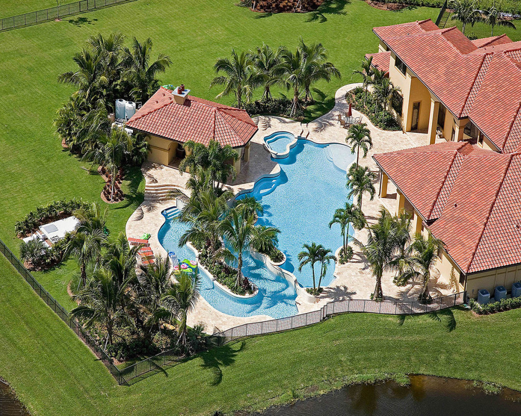 Aerial view of luxury villa with large pool, palm trees, and manicured lawn, showcasing opulent outdoor living.