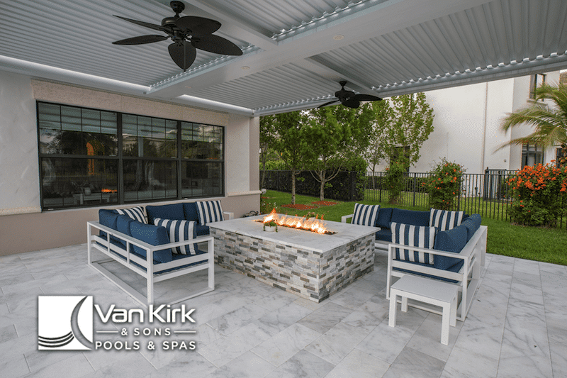 Modern outdoor patio with fire pit, blue-striped seating in lotus edge, and Van Kirk & Sons Pools & Spas logo.