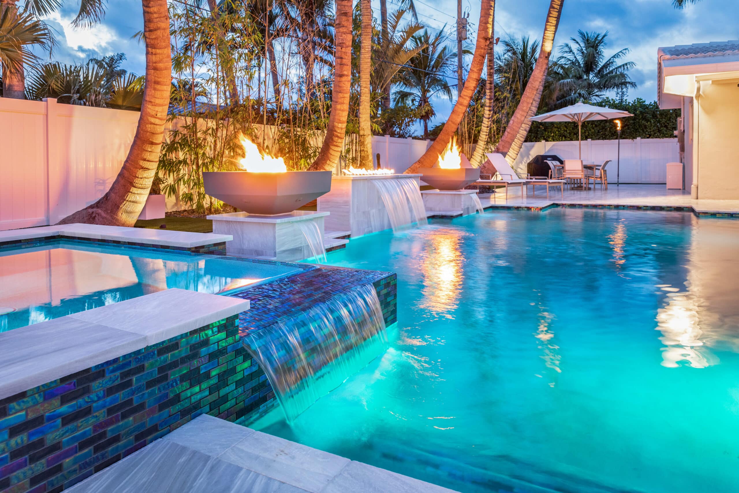 Luxurious Palm Beach Gardens, Florida backyard pool with waterfalls, fire features, and tropical palm trees at dusk.