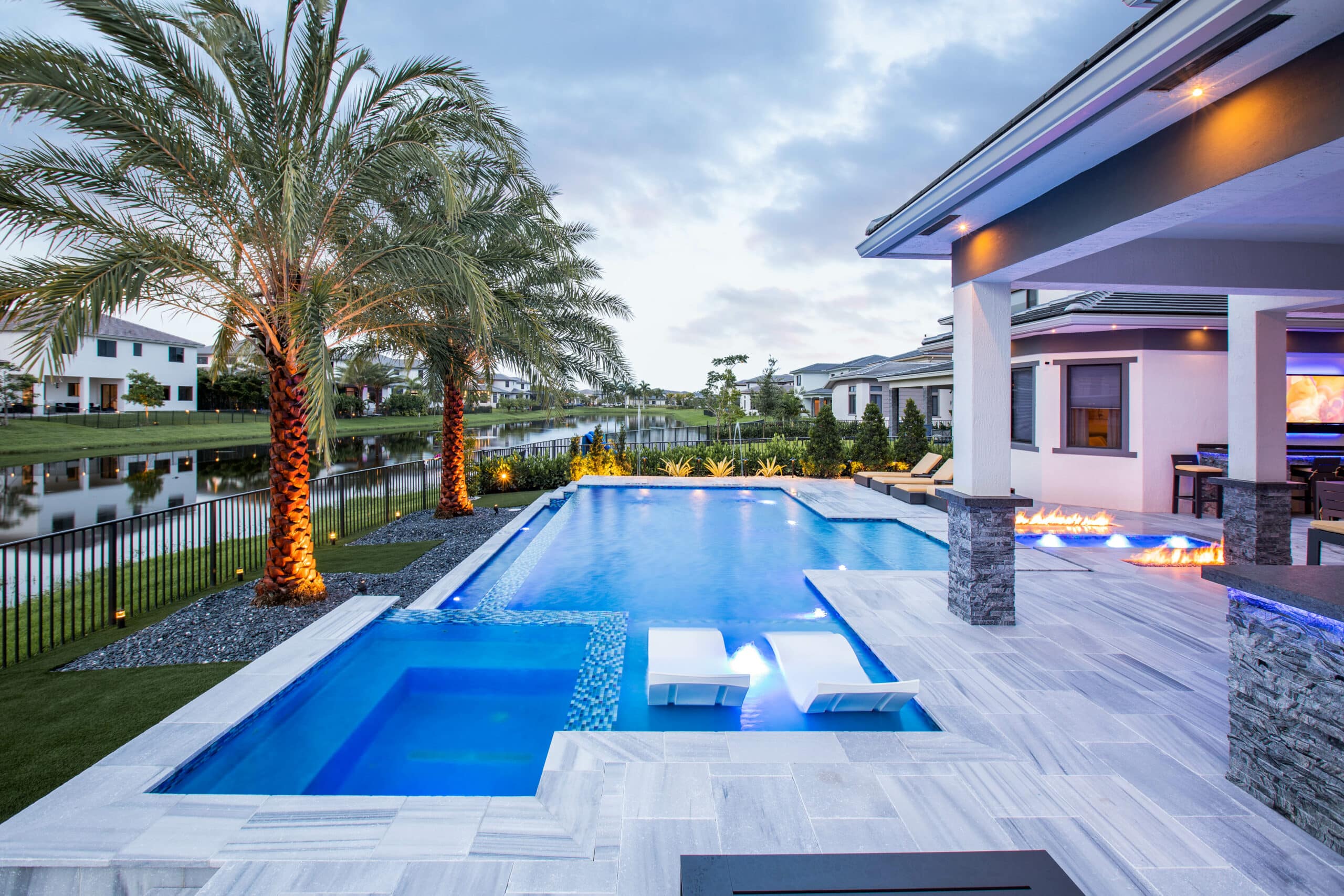 Luxury backyard oasis with palm trees, illuminated pool, fire features, and modern patio overlooking canal houses in lotus edge