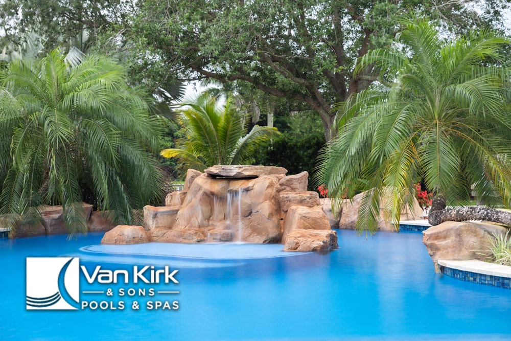 Luxurious pool with waterfall and palm trees by Van Kirk & Sons Pools & Spas, set in a tropical outdoor setting.