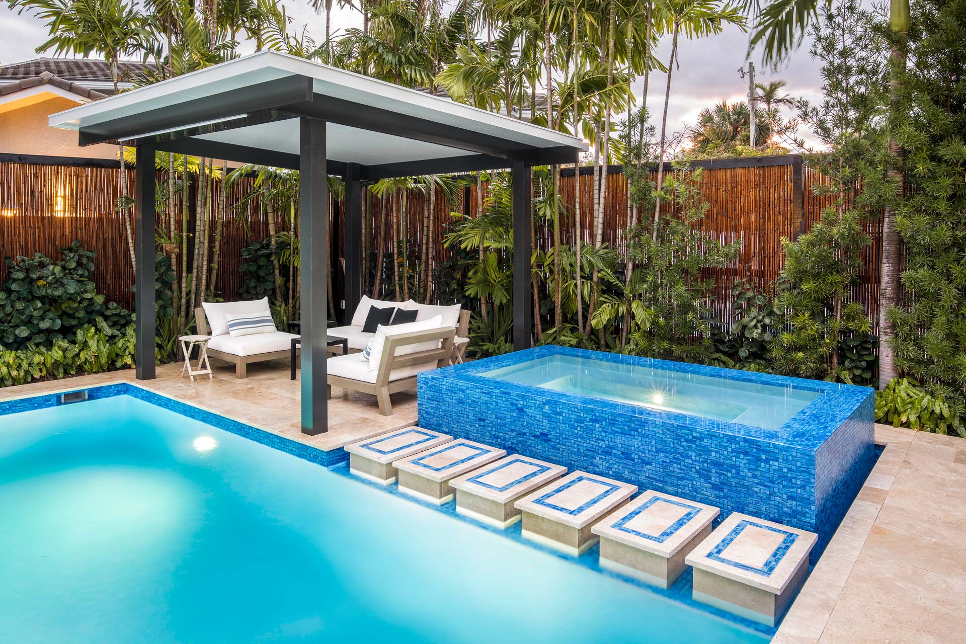 Modern backyard oasis with a blue spa, pool, swimming pool steps, and stylish shaded seating area surrounded by lush greenery.