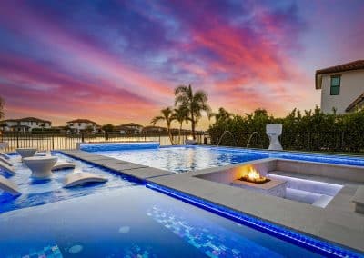 Modern pool that has a sun shelf with lounge chairs and swimming pool steps, with fire pit and palm trees at sunset, luxurious ambiance with colorful sky reflections.