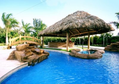 A tropical lagoon pool by Van Kirk Pools, featuring a lazy river, custom rock waterfall, wooden bridge, in-pool spa, and a large tiki hut bar.