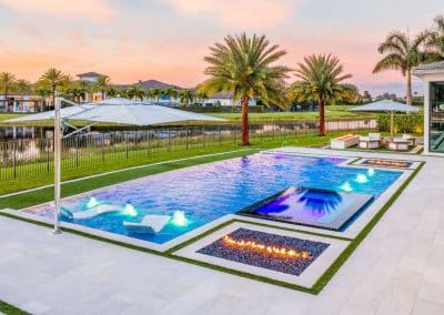 A luxurious Boca Raton backyard by Van Kirk Pools featuring a modern rectangular pool with an illuminated sun shelf, an infinity-edge spa, and a built-in fire pit at sunset.