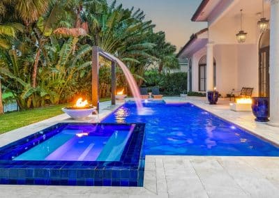 A classic linear pool at twilight with a raised blue tile spa, a high-arch waterfall, and multiple fire features.