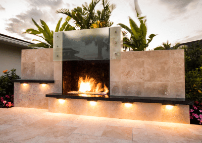 A custom outdoor fireplace with a glass backdrop, LED lighting, and travertine stone in Fort Lauderdale.