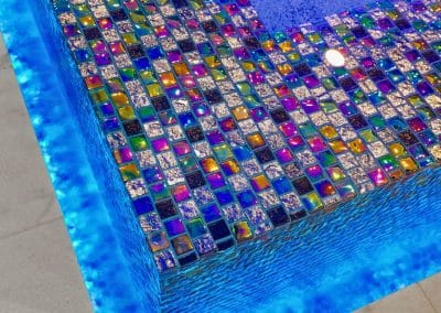 A close-up of a spa's perimeter overflow edge, featuring iridescent glass mosaic tile and a glowing blue LED light.