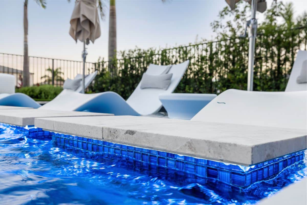 A close-up of a poolside stepping stone with blue mosaic tile and hidden LED lighting, creating a glowing effect on the water.