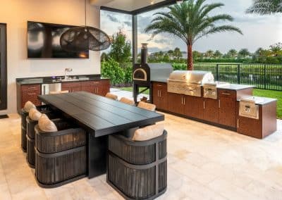 A luxury covered outdoor kitchen with a gas grill and pizza oven, next to a large dining table with a TV on the wall.
