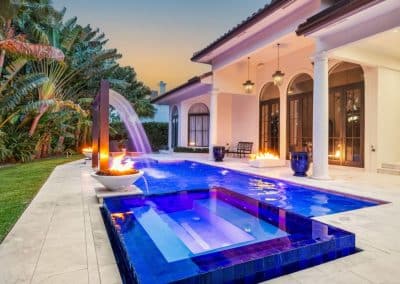 A luxury pool at sunset with a raised blue-tiled spa, a tall arching waterfall, and multiple fire pit features.
