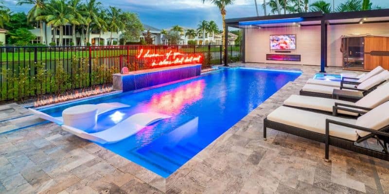 A modern Miami pool at night with a custom neon sign, a fire feature, a sun shelf, and a cabana with a TV and sauna.