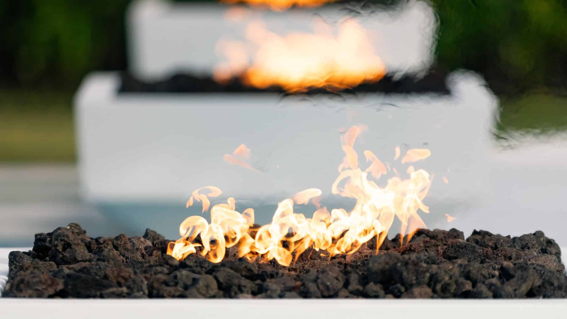 An artistic close-up shot of a modern, white fire pit with flames dancing over a bed of black lava rock.