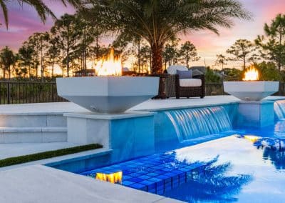 A modern raised pool wall at sunset with three square fire bowls and sheer descent waterfalls cascading into the pool.