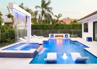 A modern geometric pool with a white rain curtain arch, a raised spa, a sun shelf with loungers, and a fireplace.
