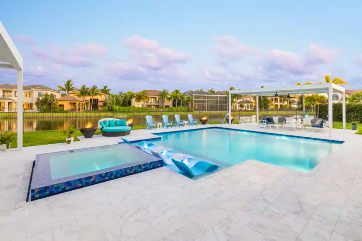 A modern pool with unique waterfall loungers, a raised spa, and a white pergola at a waterfront Boynton Beach home.