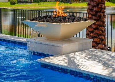 A modern, square fire and water bowl with lava rock, spilling water from a scupper into a blue mosaic tile swimming pool.