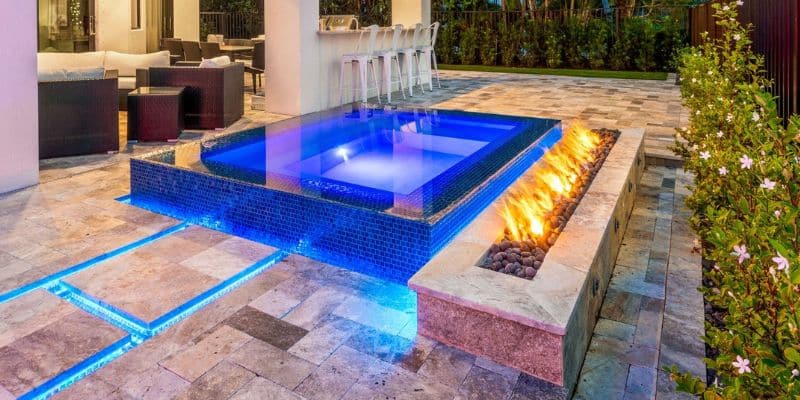 A raised, perimeter overflow spa with blue LED lighting next to a long, linear travertine fire pit on a patio at night.